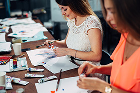 two women painting together with watercolors on paper in an art studio surrounded by paint tubes and brushes